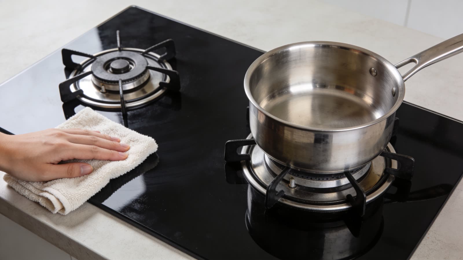 A hand cleaning a black glass top stove with a white microfiber cloth beside a metal pot.